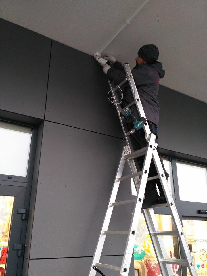 Mężczyzna w czapce i rękawicach montuje kamerę monitoringu na suficie, stojąc na aluminiowej drabinie, z wiertarką przymocowaną do drabiny.