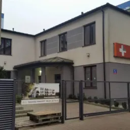 Dwupiętrowy budynek centrum medycznego Lidermed z pomarańczowym szyldem z białym krzyżem, widoczny zza metalowego ogrodzenia z reklamą sprzedaży mieszkań.