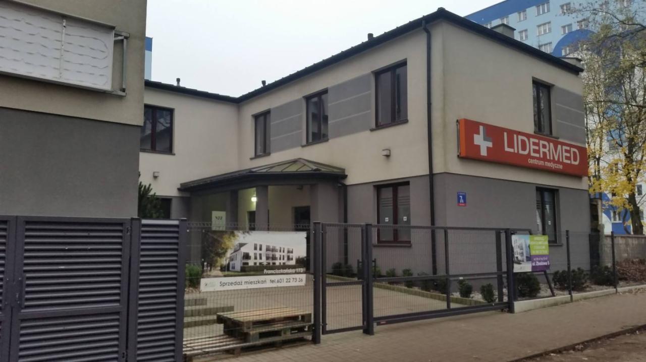Dwupiętrowy budynek centrum medycznego Lidermed z pomarańczowym szyldem z białym krzyżem, widoczny zza metalowego ogrodzenia z reklamą sprzedaży mieszkań.