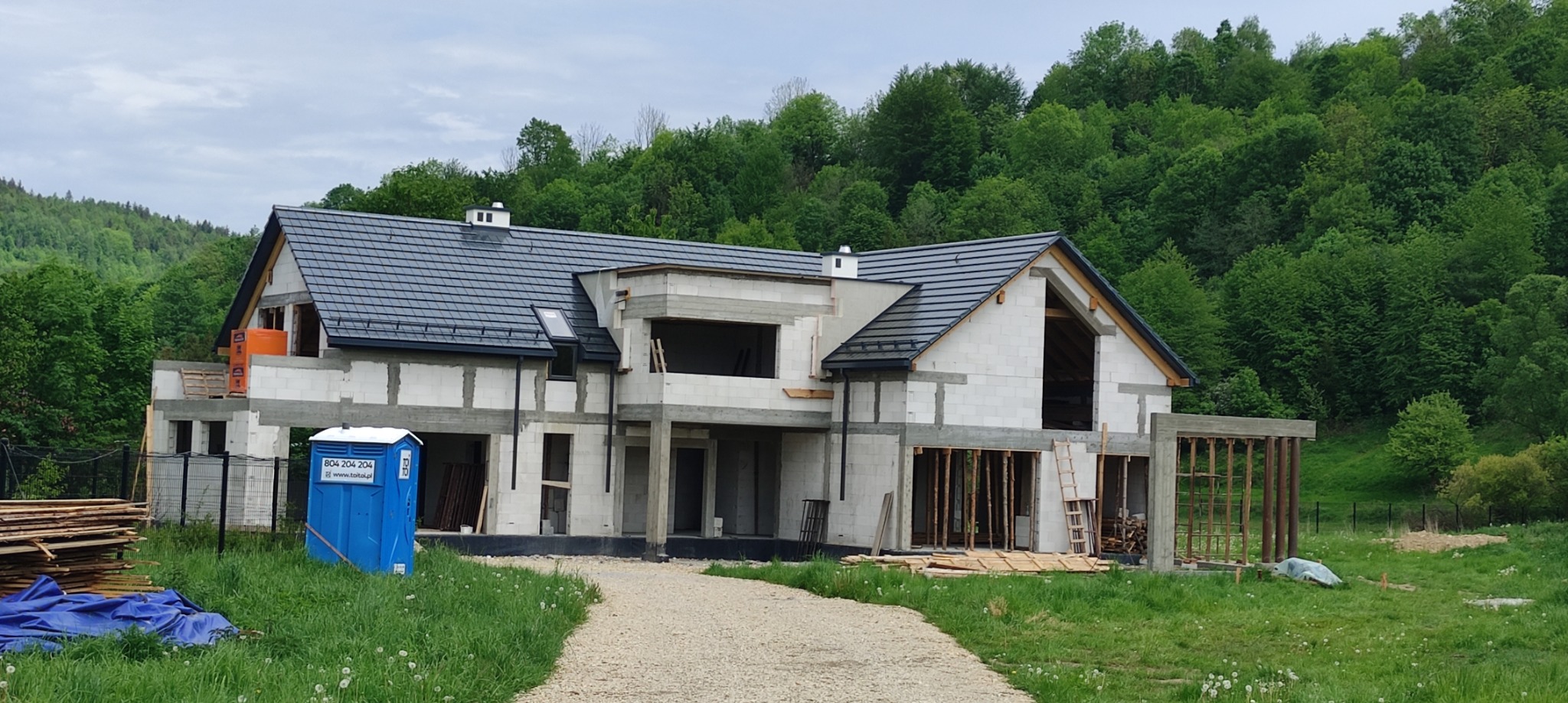 Surowy stan dwukondygnacyjnego budynku z szarym dachem w trakcie budowy, widoczny przenośny sanitariat i las w tle.