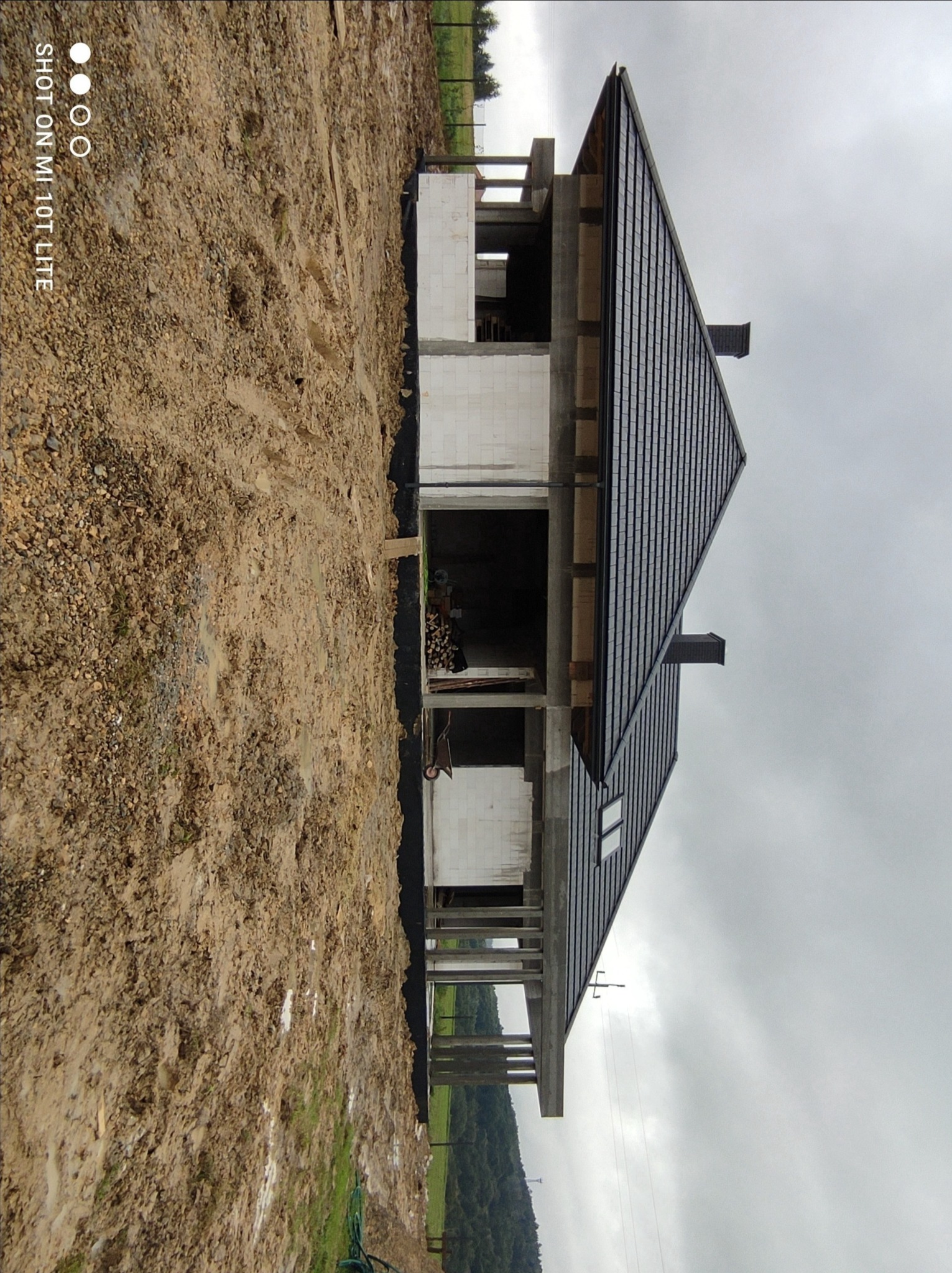 Surowy stan domu w trakcie budowy z białego pustaka, z widocznym dachem pokrytym czarną dachówką, na grząskim terenie i pochmurnym niebie.