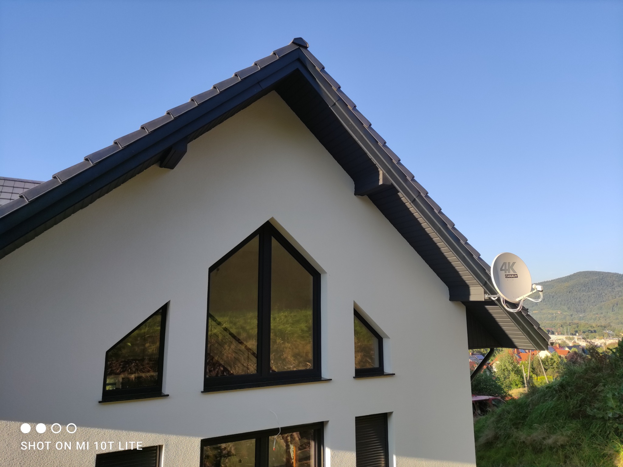 Fragment nowoczesnego domu z białymi ścianami, ciemną dachówką i oknami o nietypowych kształtach trójkątnych, antena satelitarna na dachu, w tle zieleń i góry.