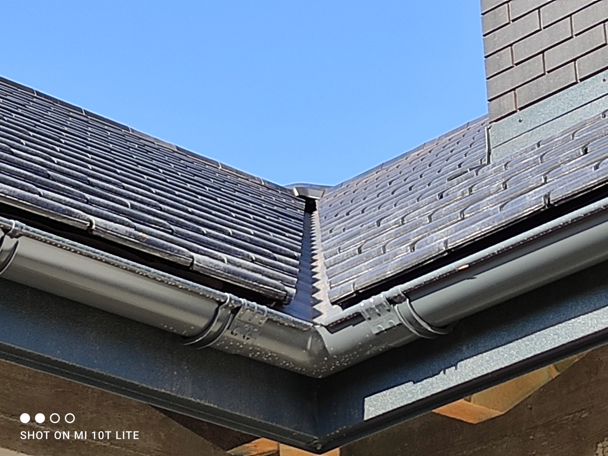 Szczegółowe ujęcie dachu z ciemnej dachówki ceramicznej i szarej rynny, widoczne mocowania rynny, fragment komina w tle na tle błękitnego nieba.