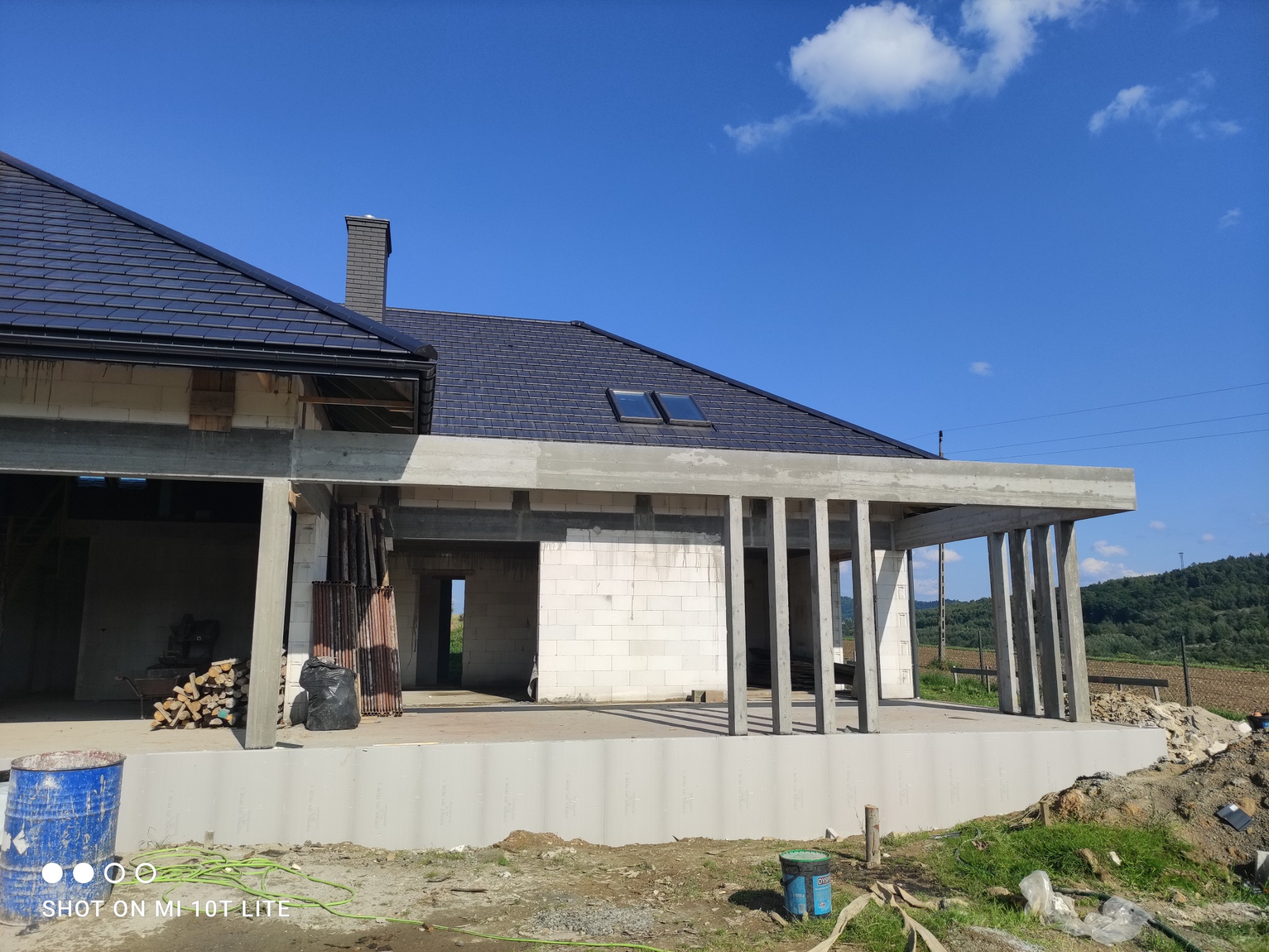 Wznoszony dom murowany w stanie surowym otwartym z ciemną dachówką, widoczne bloczki, betonowe słupy i elementy konstrukcyjne na tle błękitnego nieba.