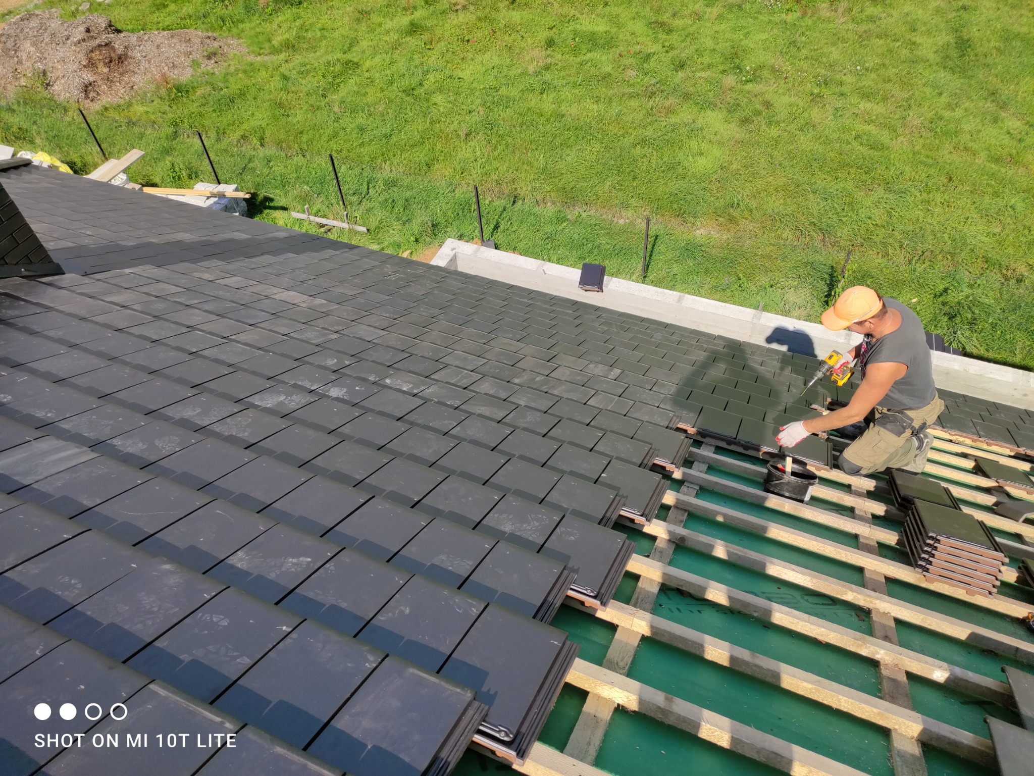 Montaż czarnej dachówki na drewnianej konstrukcji dachu, widok z góry na dekarza z wkrętarką na tle zielonej trawy.