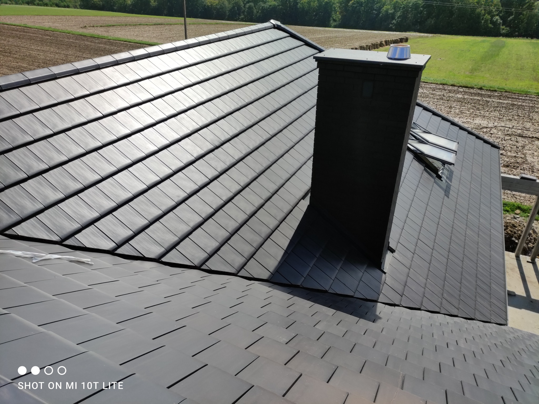 Nowoczesny dach z grafitowej dachówki ceramicznej z kominem i oknami dachowymi, widok z góry na tle krajobrazu.