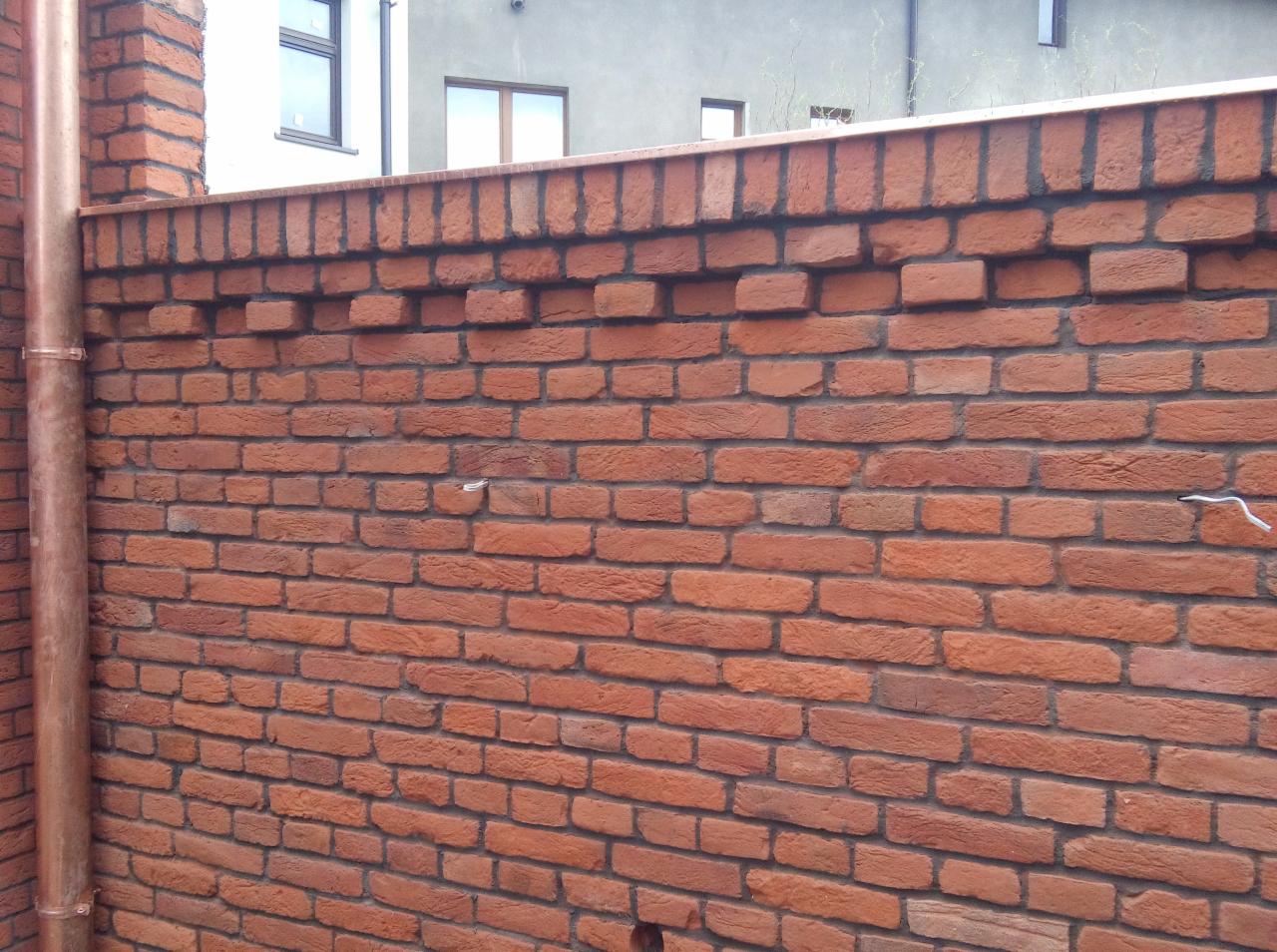 Fragment muru z czerwonej cegły z wystającymi elementami i metalowym wykończeniem, widoczny fragment rynny i okna budynku w tle.