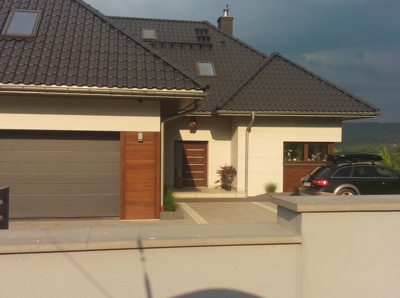 Nowoczesny dom z szarą dachówką, garażem z segmentową bramą i drewnianymi akcentami elewacji, samochód osobowy z bagażnikiem dachowym zaparkowany na podjeździe.