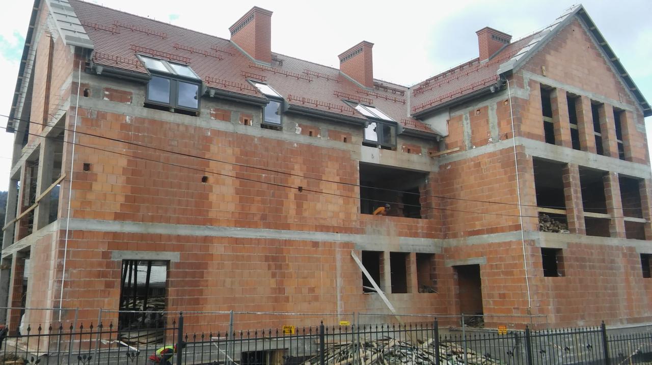 Budowa dwupiętrowego domu z czerwonej cegły z widocznymi oknami dachowymi i kominami, zabezpieczonego czarnym metalowym ogrodzeniem.