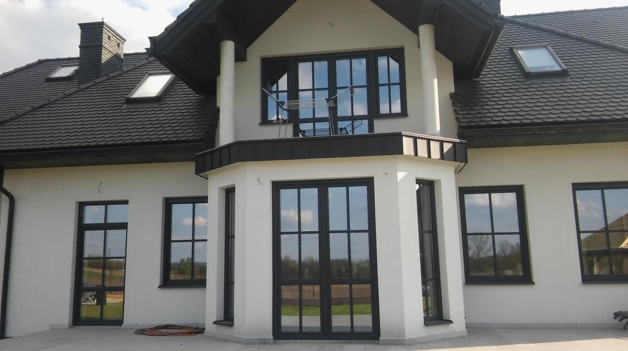 Elewacja dwukondygnacyjnego domu z białym tynkiem, czarną stolarką okienną i dachem z dachówki ceramicznej; na balkonie widoczna suszarka na pranie.