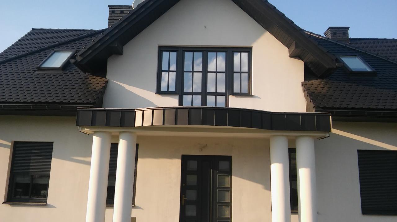 Elewacja dwukondygnacyjnego domu z białym tynkiem, ciemną dachówką i stolarką okienną, widoczne okna dachowe, kolumny przy wejściu i rolety zewnętrzne.