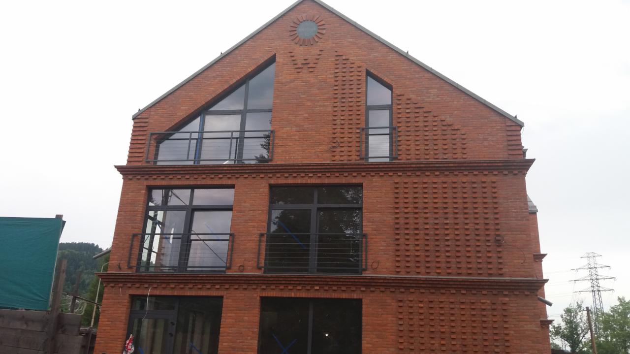 Trzykondygnacyjny budynek z czerwonej cegły z oknami o czarnych ramach i balkonami z metalowymi balustradami, widok z zewnątrz na tle pochmurnego nieba i zieleni.