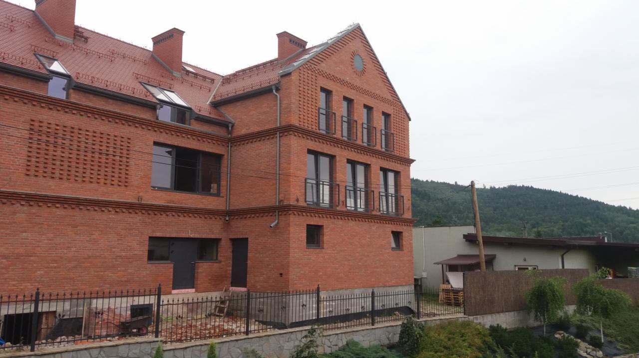 Dwukondygnacyjny budynek z czerwonej cegły z balkonami i oknami w czarnych ramach, widok z zewnątrz, w tle zalesione wzgórze.