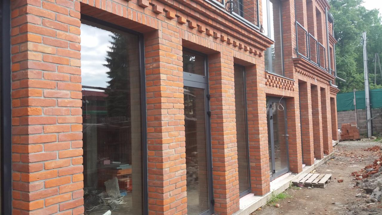 Elewacja budynku z czerwonej cegły z dużymi oknami w ciemnych ramach, widoczne elementy dekoracyjne z cegły, teren wokół budynku w trakcie prac wykończeniowych.