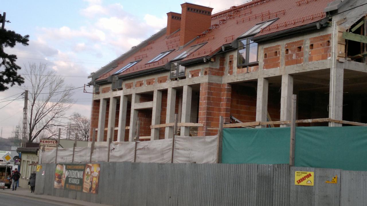 Budowa domu w stanie surowym otwartym, widoczne ceglane ściany, betonowe słupy i czerwona dachówka na dachu z oknami dachowymi, zabezpieczone ogrodzeniem z blachy falistej.