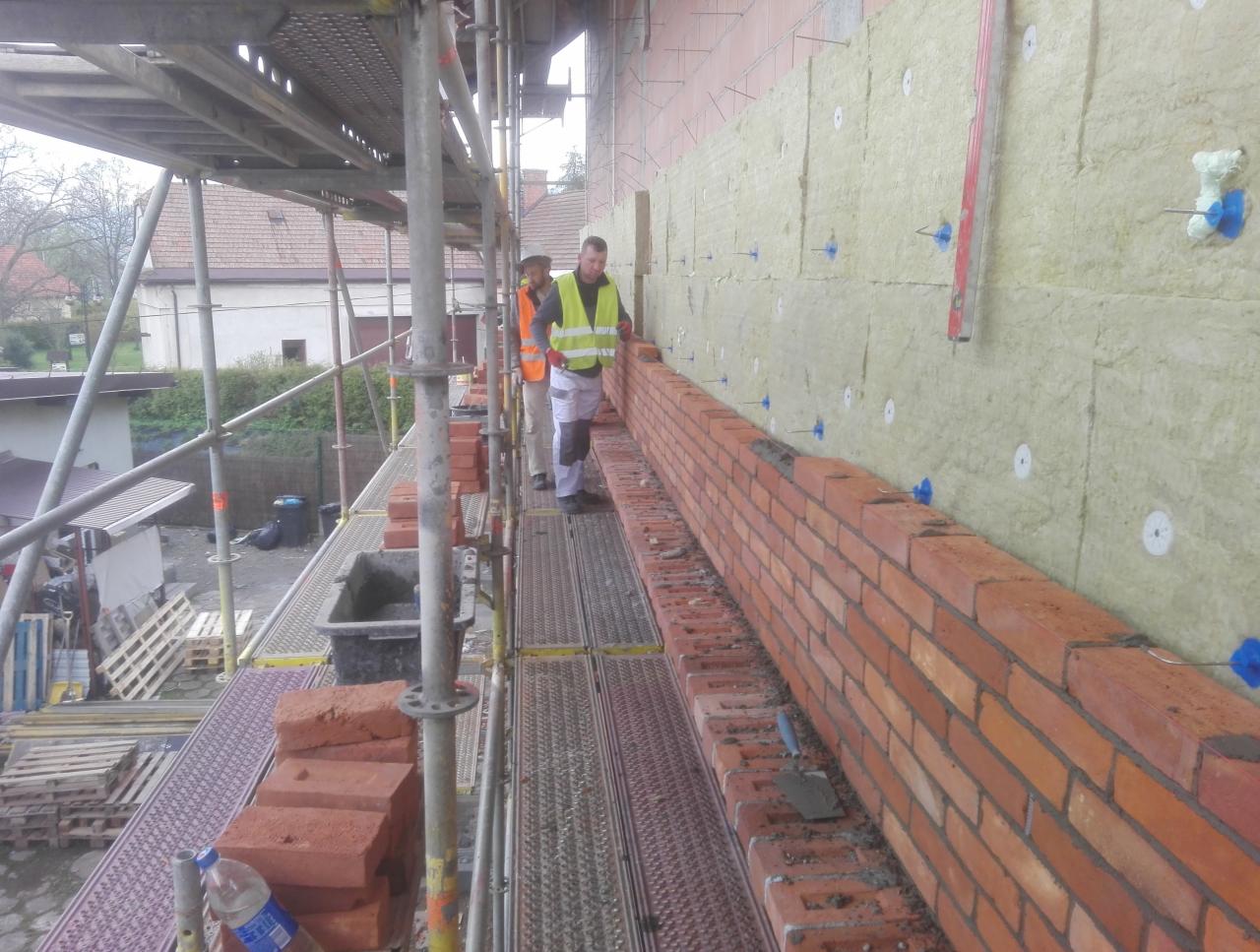 Prace murarskie na rusztowaniu: układanie czerwonej cegły na ścianie z izolacją, widoczni dwaj pracownicy w kamizelkach odblaskowych.