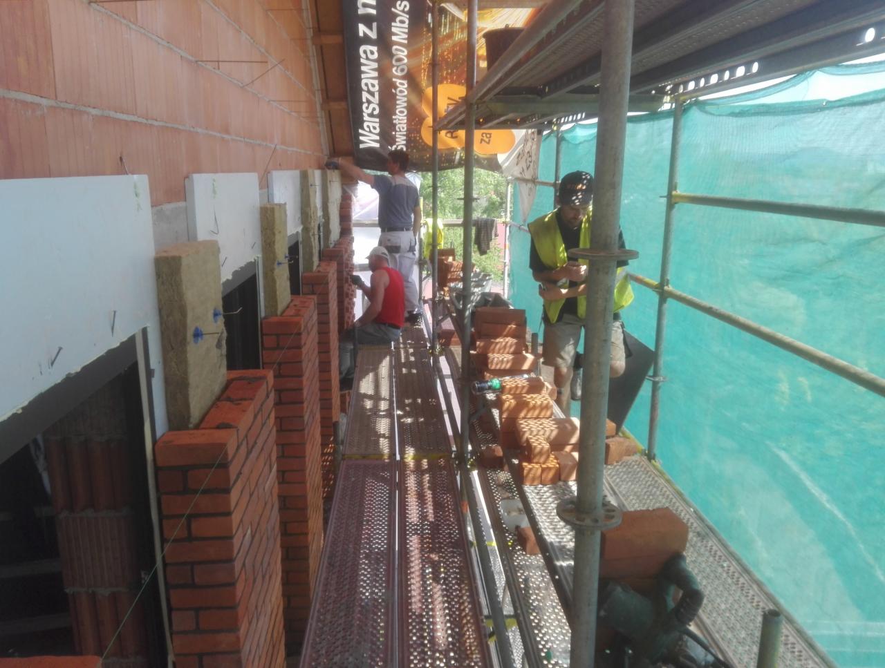 Budowa ściany zewnętrznej z cegły klinkierowej, widoczne rusztowanie, ocieplenie między ścianą a cegłą, dwóch pracowników muruje, trzeci pracuje na górze, reklama światłowodu na budynku.