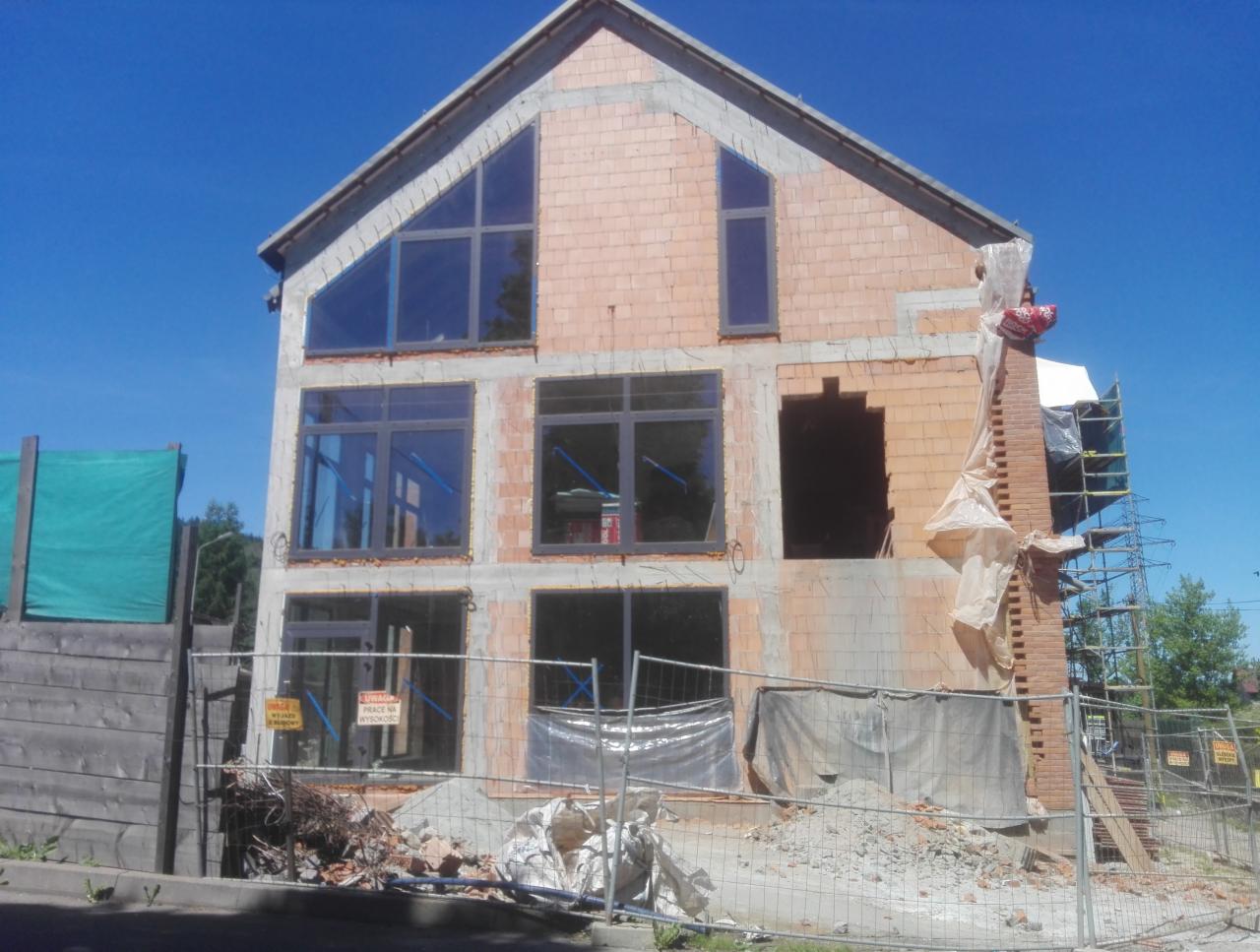 Wznoszenie dwukondygnacyjnego budynku z cegły z dużymi oknami, widoczny częściowo ukończony komin z rusztowaniem i gruz przed budynkiem.