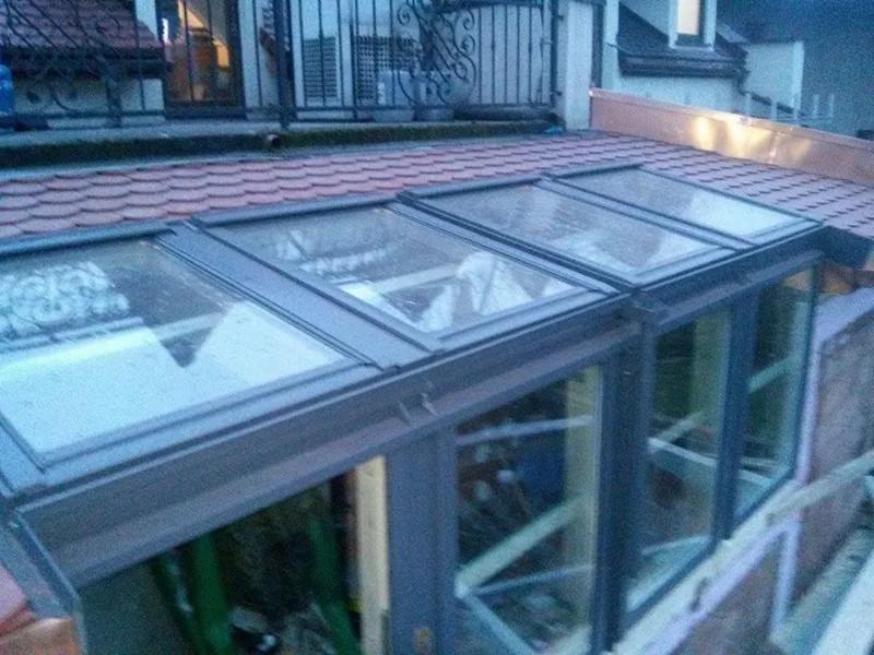 Rozbudowa domu z przeszklonym dachem i widocznymi elementami konstrukcyjnymi w trakcie budowy, czerwona dachówka w tle.