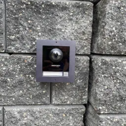 Zewnętrzny panel wideodomofonu Hikvision z kamerą typu rybie oko, zamontowany na szarym murze z betonowych bloczków.