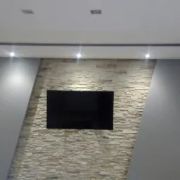 Nowoczesny salon z podwieszanym sufitem, oświetleniem punktowym i ścianą z dekoracyjnej cegły, na której wisi telewizor.