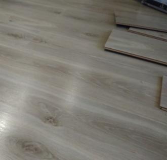 Fragment podłogi z paneli o jasnym odcieniu drewna, z widoczną teksturą i kilkoma ułożonymi panelami w rogu, sugerującymi etap montażu.