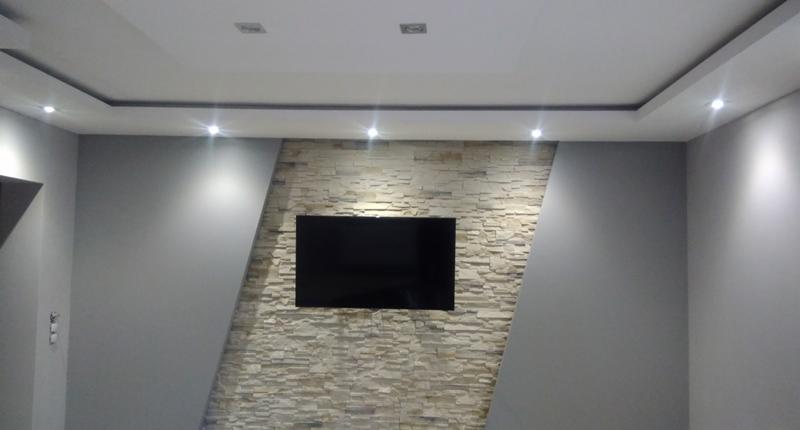 Nowoczesny salon z podwieszanym sufitem, oświetleniem punktowym i ścianą z dekoracyjnej cegły, na której wisi telewizor.