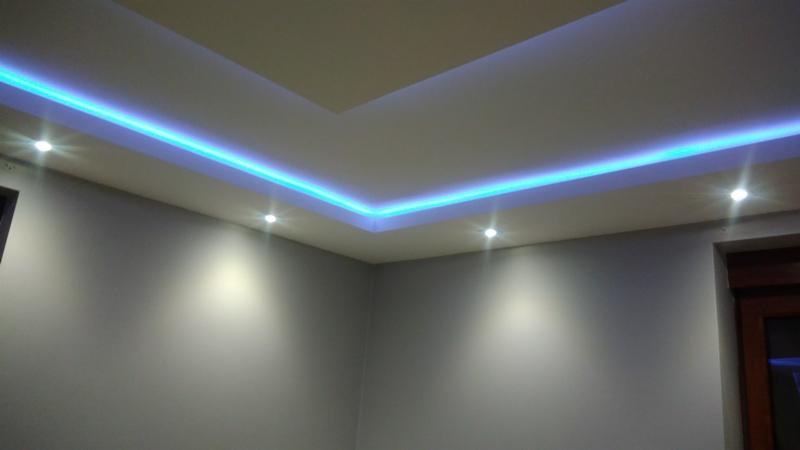 Nowoczesny sufit podwieszany z niebieskim oświetleniem LED i punktowymi lampami halogenowymi, szare ściany, widok z narożnika pokoju.