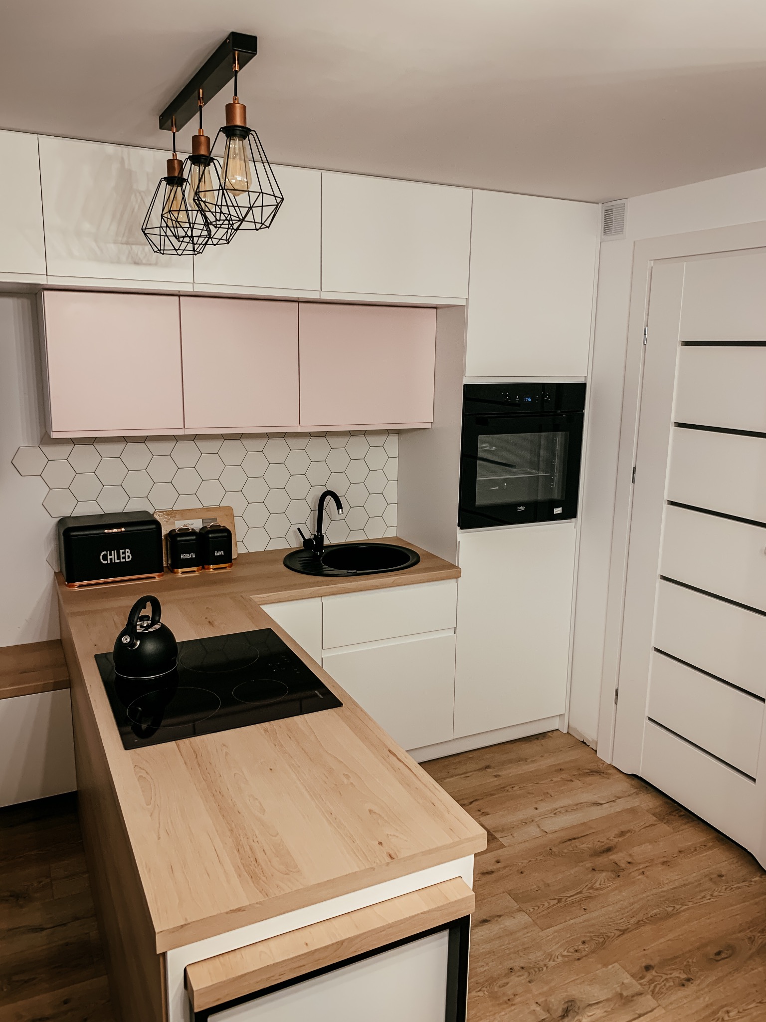 Nowoczesna kuchnia z białymi szafkami, drewnianym blatem, różowymi akcentami i heksagonalnymi płytkami na ścianie, oświetlona trzema lampami z geometrycznymi kloszami.