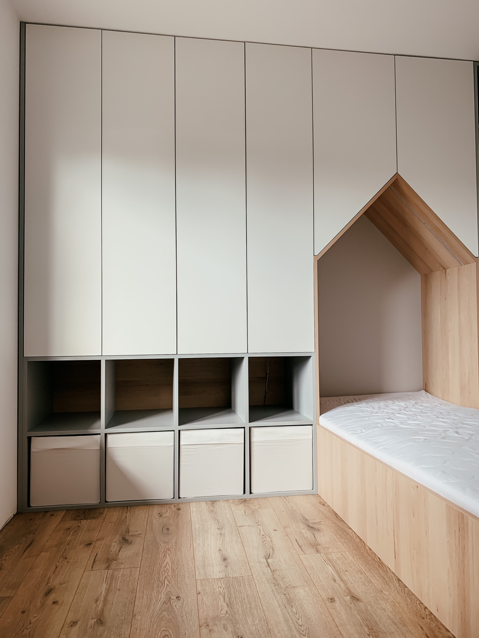 Pokój dziecięcy z zabudową: szafa z szufladami i półkami oraz łóżko w kształcie domku, wykonane z jasnego drewna i szarej płyty.