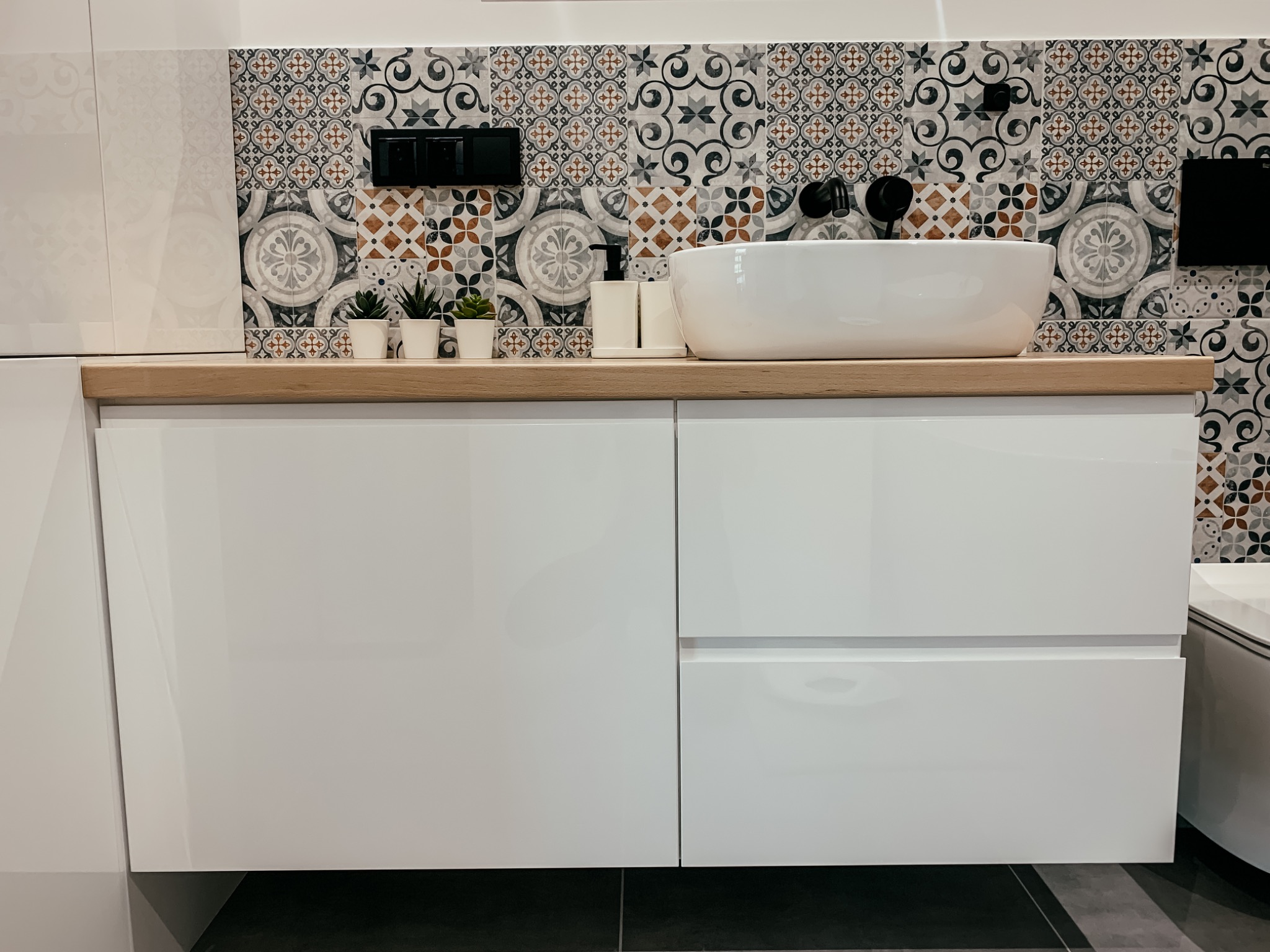 Biała, błyszcząca szafka pod umywalkę z drewnianym blatem, na którym stoją małe sukulenty w białych doniczkach, biały dozownik do mydła oraz owalna umywalka nablatowa. Tło stanowi ściana...