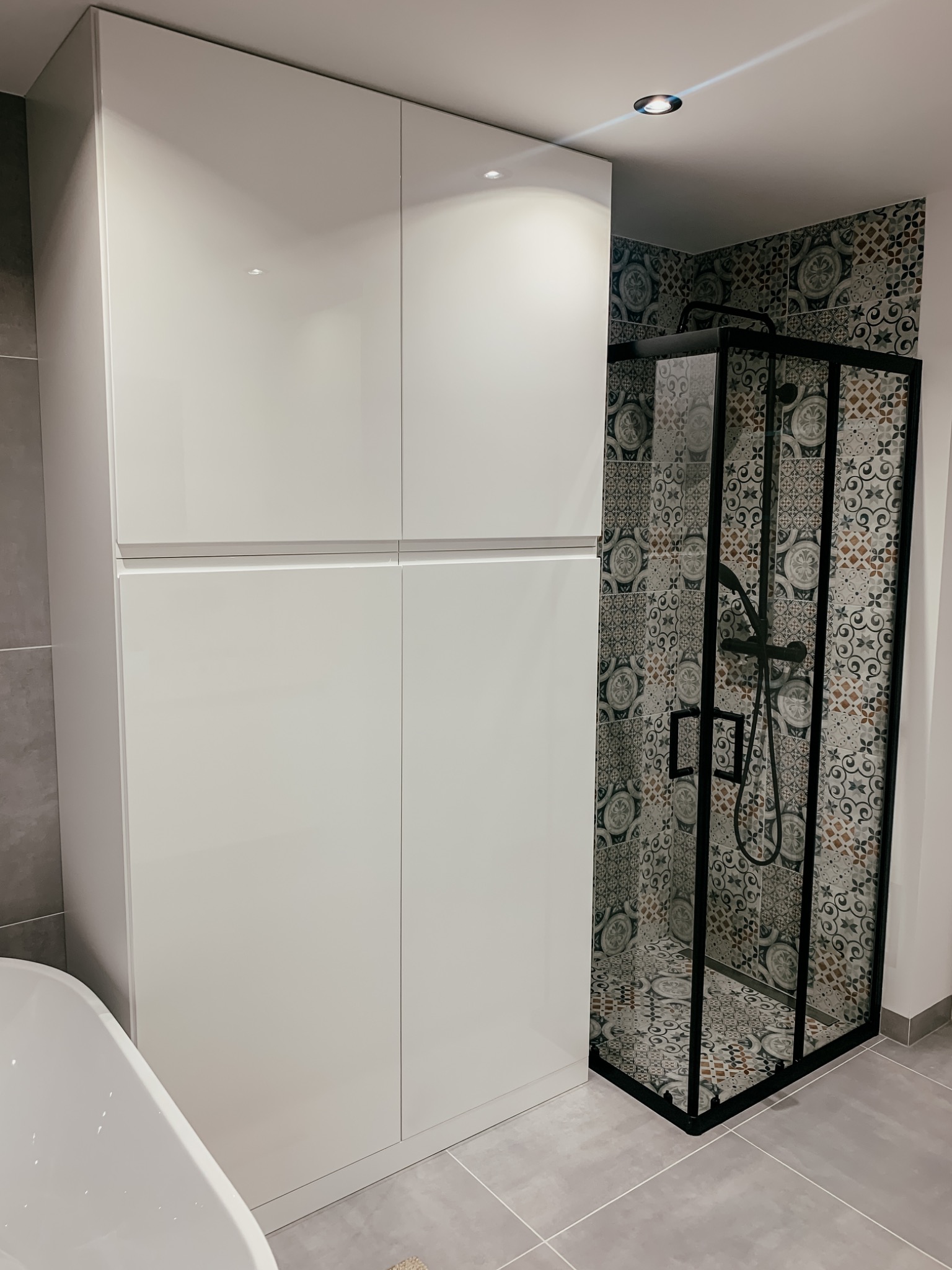 Biała, wysoka szafa łazienkowa z połyskiem, sąsiadująca z kabiną prysznicową z czarnymi profilami i wzorzystymi płytkami w tle.
