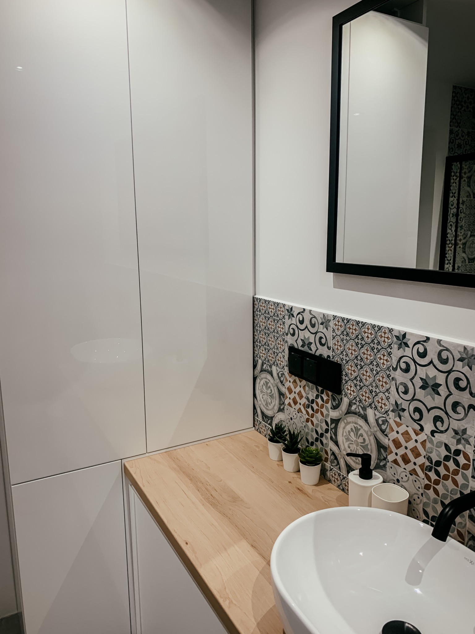 Nowoczesna łazienka z białą szafką, drewnianym blatem, dekoracyjnymi płytkami i okrągłą umywalką nablatową z czarną armaturą.