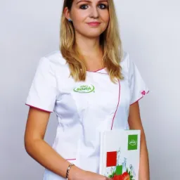 Portret uśmiechniętej kobiety w białym uniformie z logo dietetyka, trzymającej broszurę z motywem truskawki i kropli wody.