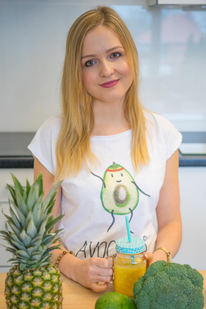 Uśmiechnięta blondynka w białej koszulce z awokado trzyma słoik z pomarańczowym smoothie, obok ananas i brokuł na drewnianym blacie w kuchni.