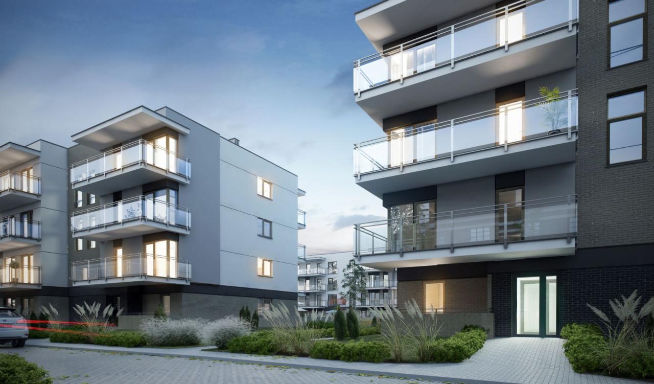 Nowoczesne budynki apartamentowe z balkonami wykończonymi szkłem, otoczone niską roślinnością ozdobną i brukowanym chodnikiem, z widocznymi śladami świateł przejeżdżającego samochodu po lewej stronie.