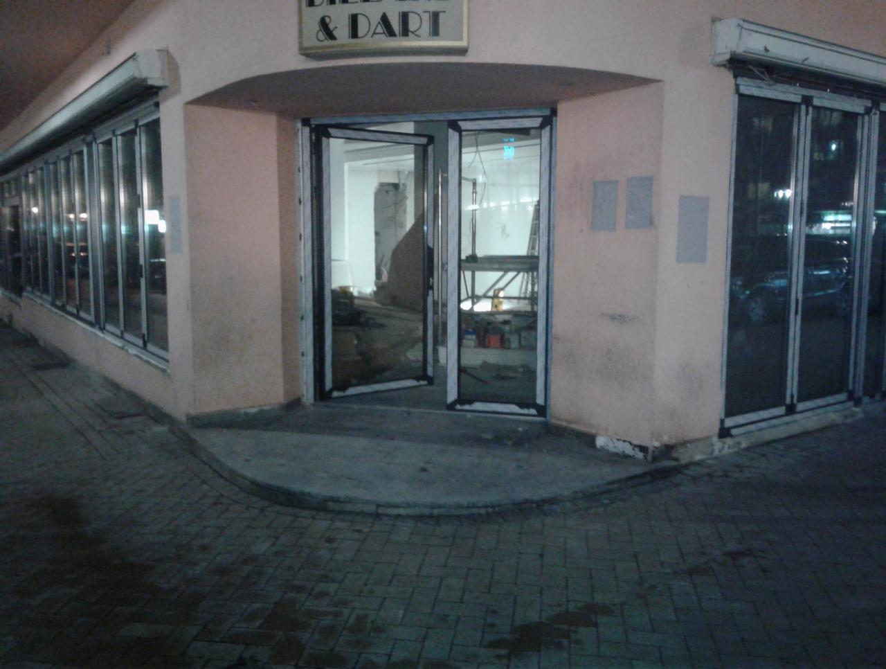 Nocne ujęcie wejścia do lokalu z otwartymi, przeszklonymi drzwiami i witrynami okiennymi, widoczne rusztowanie wewnątrz, oświetlenie uliczne.