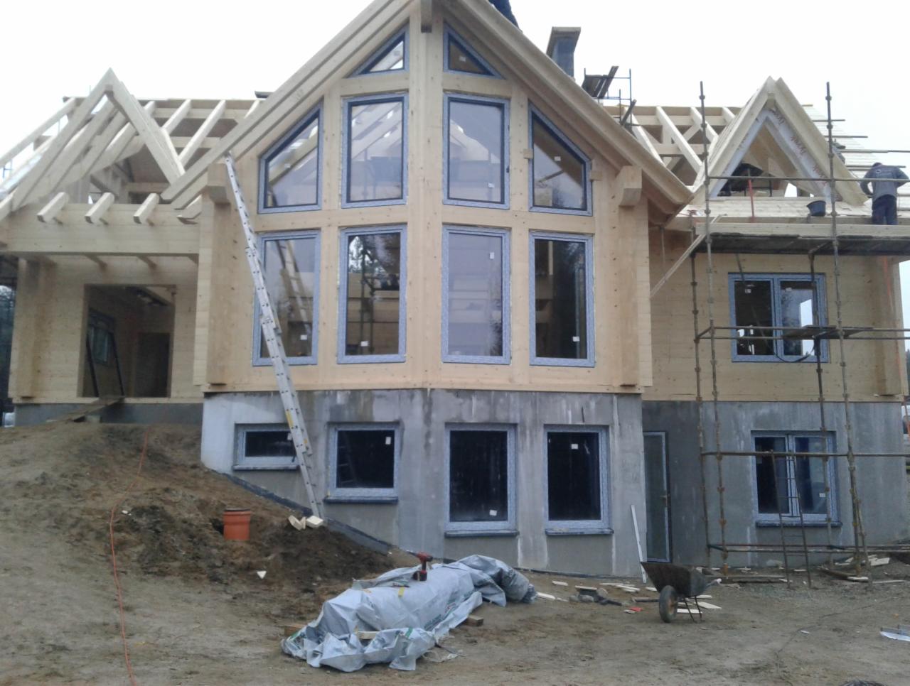 Surowy stan domu z drewna podczas budowy, z zamontowanymi oknami z szarymi ramami, widoczne rusztowania i materiały budowlane.