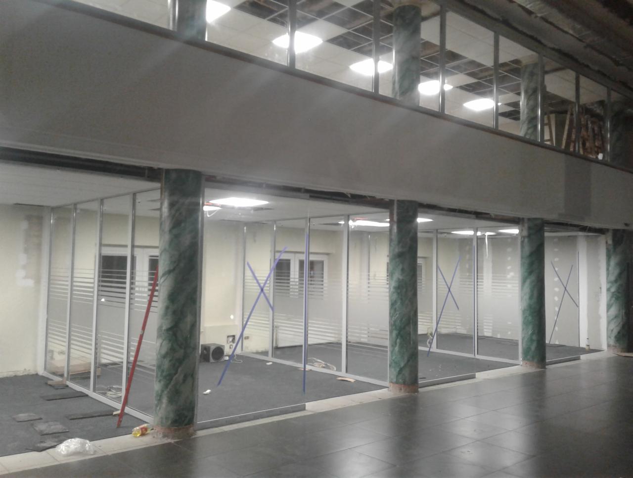 Montaż szklanych ścianek działowych w biurze, widoczne marmurowe kolumny i niebieskie taśmy stabilizujące, w tle okna i sufit podwieszany.