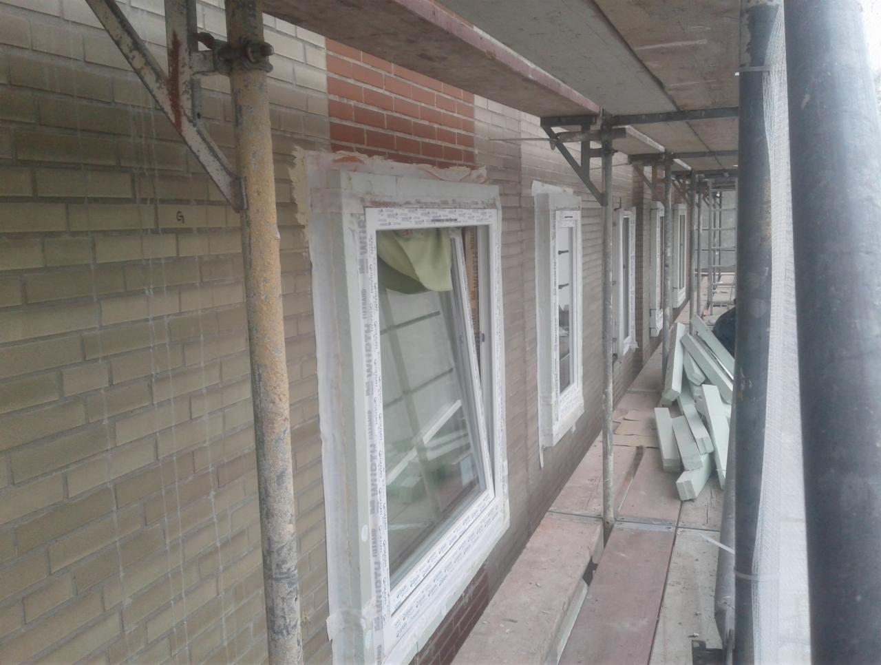 Rząd okien w trakcie montażu na tle ceglanej ściany, widoczne rusztowanie i materiały izolacyjne na podeście.