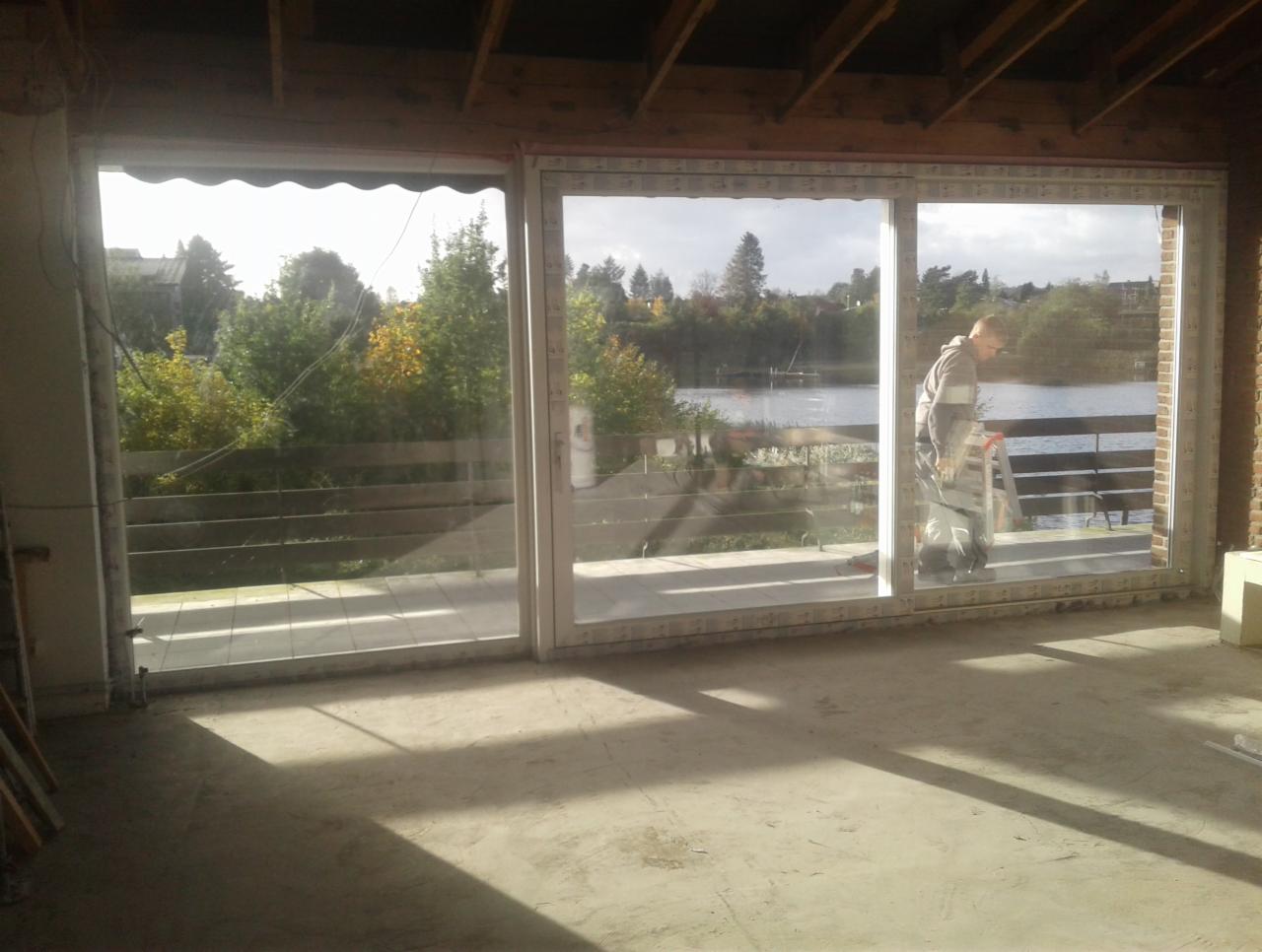 Monolityczne okno balkonowe z białymi ramami, widok na jezioro i zieleń, pracownik na drabinie w trakcie montażu, surowy beton na podłodze, widoczne drewniane belki stropowe.