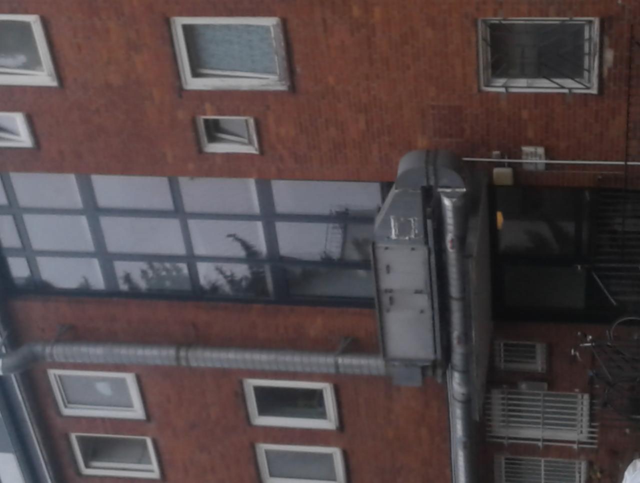 Widok z góry na budynek z cegły z różnymi oknami, w tym oknami kratowymi i dużym oknem z podziałami, z widoczną rurą spustową i elementami wentylacji.