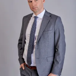 Portret mężczyzny w szarym garniturze, białej koszuli i wzorzystym krawacie, z ręką w kieszeni, pozującego na szarym tle.