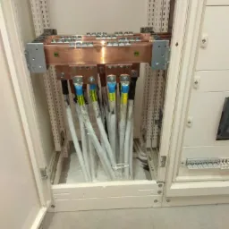 Wnętrze szafy elektrycznej z miedzianymi szynami zbiorczymi i podłączonymi kablami zabezpieczonymi folią, widoczne oznaczenia na kablach.