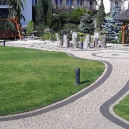 Ogród z ścieżką z kostki brukowej, altaną z trzcinowym dachem i drewnianą huśtawką w tle.