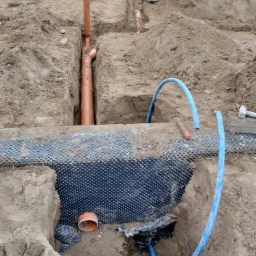 Wykop pod fundamenty z widocznymi rurami drenażowymi, folią kubełkową i elementami szalunku fundamentowego w tle. Na pierwszym planie niebieski przewód. Widoczny fragment logo w prawym dolnym rogu.