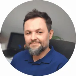 Portret uśmiechniętego mężczyzny z siwiejącą brodą w niebieskiej koszulce polo, siedzącego na krześle biurowym z rozmytym laptopem w tle, w okrągłym wykadrowaniu.