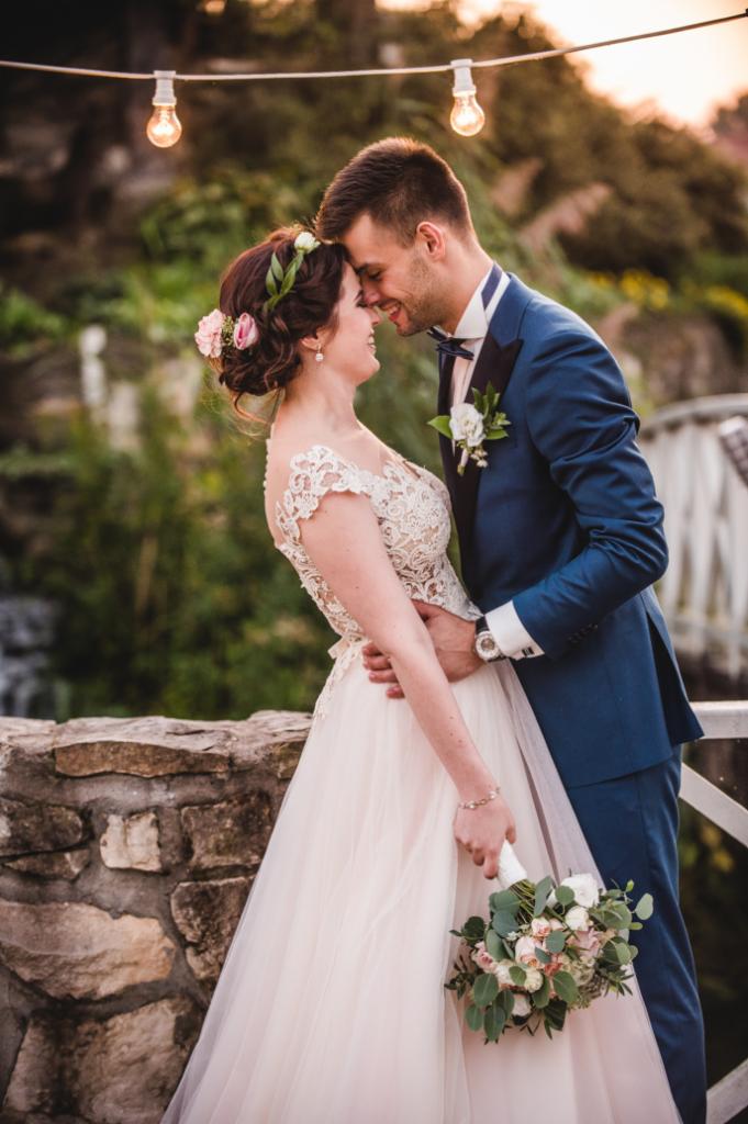 Uśmiechnięta para młoda, on w granatowym garniturze, ona w koronkowej sukni, stojąca na kamiennym murku z bukietem, oświetlona dekoracyjnymi żarówkami, z zielenią w tle.