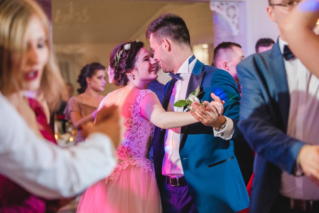 Para młoda tańczy podczas wesela, panna młoda w różowej sukni z koronkową górą i wiankiem, pan młody w niebieskim smokingu z białą butonierką, wokół inni goście weselni.