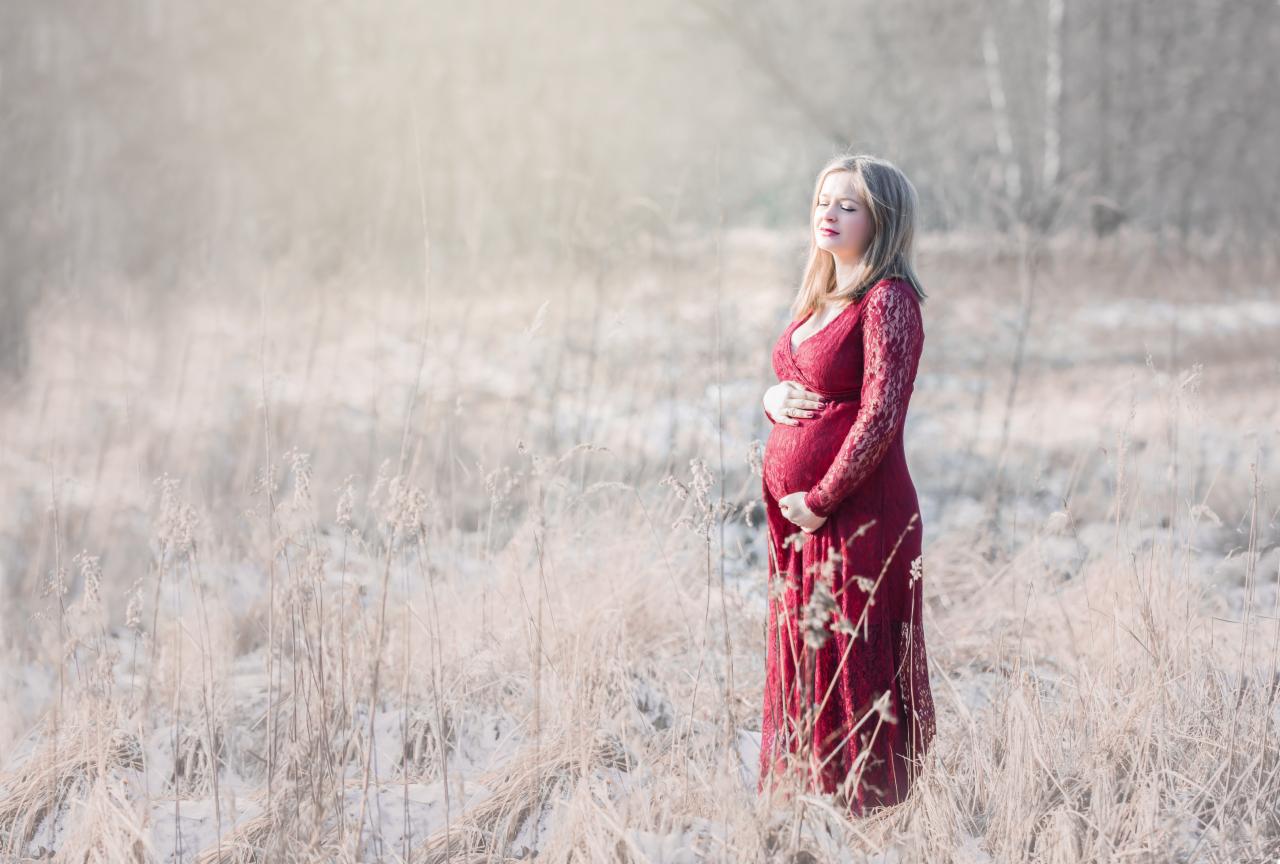 Kobieta w czerwonej koronkowej sukni ciążowej, z zamkniętymi oczami, trzymająca dłonie na brzuchu, pozuje na tle łąki w zimowej scenerii.